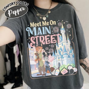 May include: A dark gray t-shirt with the words "Meet Me On Main Street" in colorful text. The graphic features cartoon characters, a castle, fireworks, and other theme park elements. The shirt has the words "Comfort Colors" and "Pepper" on the top left.