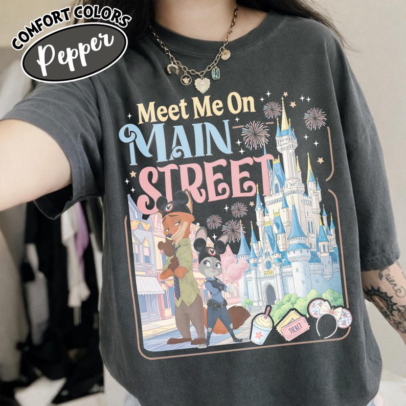 May include: A dark gray t-shirt with the words "Meet Me On Main Street" in colorful text. The graphic features cartoon characters, a castle, fireworks, and other theme park elements. The shirt has the words "Comfort Colors" and "Pepper" on the top left.