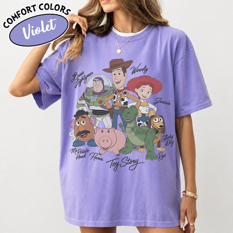 May include: Violet Comfort Colors t-shirt featuring a graphic of Toy Story characters including Woody, Buzz Lightyear, and Jessie. The shirt has a crew neck and short sleeves. The text "Toy Story" is printed below the characters.
