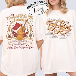 May include: An ivory t-shirt featuring two graphic designs. The back displays a red cowboy hat, acoustic guitar, roses, a horseshoe, and a steer skull with the text Cowgirl Like Me and Eyes full of stars... Takes One to Know One. The front shows the text it's a Toy Story Baby, just say yes in rope-style lettering.