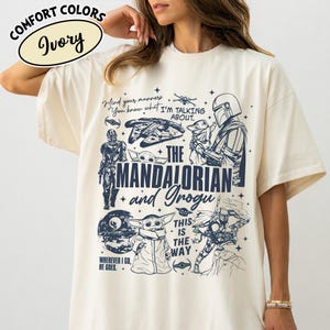 Op de afbeelding: Ivoor kleurig t-shirt met een blauw grafisch ontwerp met personages en citaten uit "The Mandalorian". Het ontwerp bevat de tekst "The Mandalorian and Grogu" en "This is the Way".