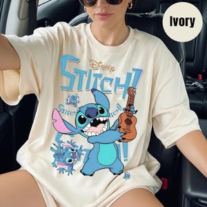 Pu&ograve; includere: T-shirt color avorio con il personaggio Disney Stitch che tiene un ukulele. Il design include la parola "Stitch!" in blu, insieme ad altre illustrazioni di Stitch. La maglietta &egrave; un capo casual.