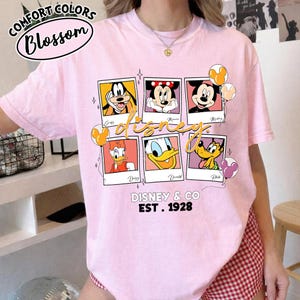 Op de afbeelding: Zachtroze T-shirt met een Disney-thema. Het ontwerp bevat polaroid-stijl frames met personages als Goofy, Minnie, Mickey, Daisy, Donald en Pluto. De tekst "Disney & Co Est. 1928" is eronder gedrukt.