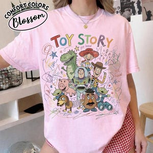 May include: Pink t-shirt featuring a colorful graphic of Toy Story characters and the words "Toy Story" in green and red. The shirt also has the Comfort Colors and Blossom logos.