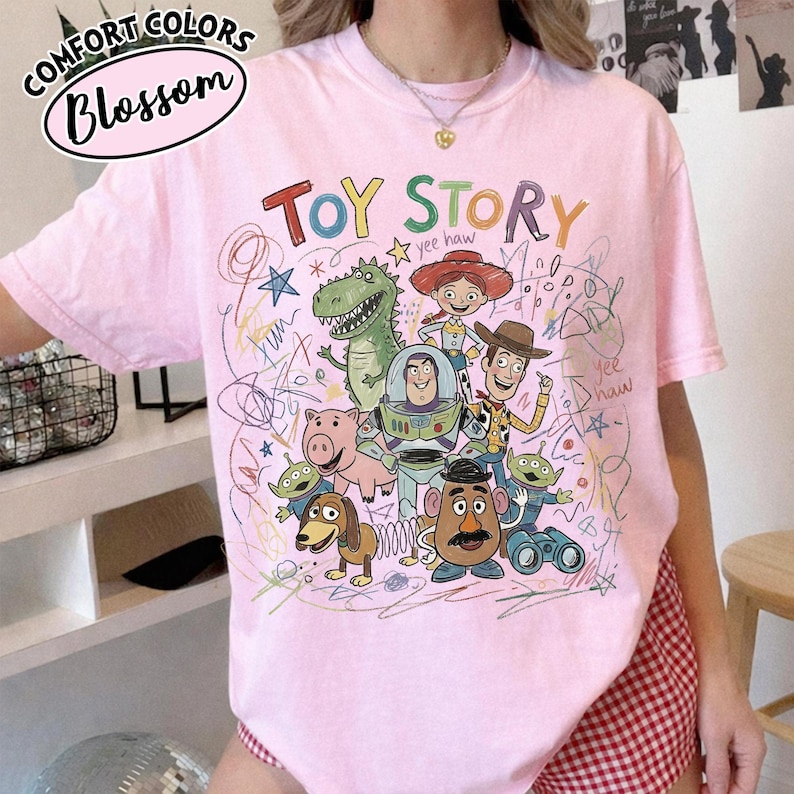 May include: Pink t-shirt featuring a colorful graphic of Toy Story characters and the words "Toy Story" in green and red. The shirt also has the Comfort Colors and Blossom logos.