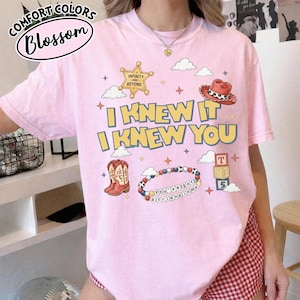 May include: A light pink t-shirt featuring a graphic print with the text "I KNEW IT I KNEW YOU" in yellow and blue. The design includes a gold sheriff star, a red cowboy hat, a pair of brown cowboy boots, a beaded bracelet, and alphabet blocks.
