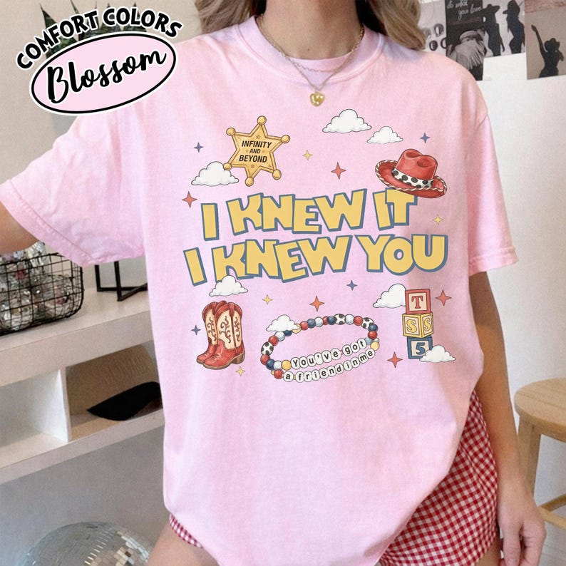May include: A light pink t-shirt featuring a graphic print with the text "I KNEW IT I KNEW YOU" in yellow and blue. The design includes a gold sheriff star, a red cowboy hat, a pair of brown cowboy boots, a beaded bracelet, and alphabet blocks.