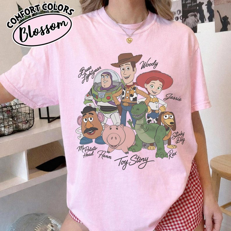 May include: A light pink t-shirt featuring a graphic of characters from the animated film "Toy Story." The design includes Buzz Lightyear, Woody, Jessie, Mr. Potato Head, Hamm, Rex, and Slinky Dog. The shirt has the text "Comfort Colors Blossom" at the top.
