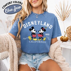 K&ouml;nnte beinhalten: Blaues Jeans-T-Shirt mit dem Wort "Disneyland" und einem Bild von Mickey und Minnie Mouse. Das Shirt enth&auml;lt auch den Text "1955 California" und die Worte "Comfort Colors Blue Jean" in einem kreisf&ouml;rmigen Design.