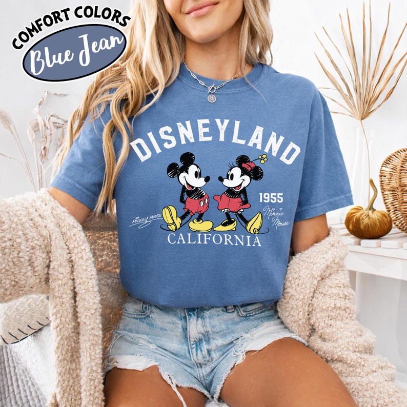 K&ouml;nnte beinhalten: Blaues Jeans-T-Shirt mit dem Wort "Disneyland" und einem Bild von Mickey und Minnie Mouse. Das Shirt enth&auml;lt auch den Text "1955 California" und die Worte "Comfort Colors Blue Jean" in einem kreisf&ouml;rmigen Design.
