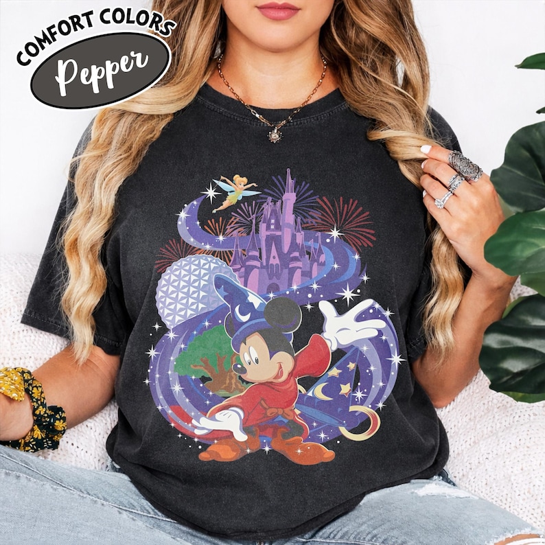 Może przedstawiać: Czarny t-shirt z kolorową grafiką przedstawiającą Myszkę Miki w stroju czarodzieja, zamek, fajerwerki i napisem "Comfort Colors Pepper". Projekt zawiera wr&oacute;żkę, globus i wirujące fioletowe tło.