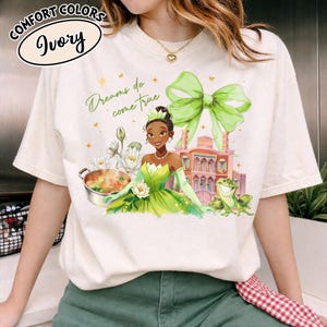 Puede incluir: Camiseta color marfil con una ilustraci&oacute;n de princesa con un vestido verde, una rana y un edificio. El texto "Dreams do come true" est&aacute; escrito en verde. La camiseta tambi&eacute;n tiene las palabras "Comfort Colors" e "Ivory" en un dise&ntilde;o circular.