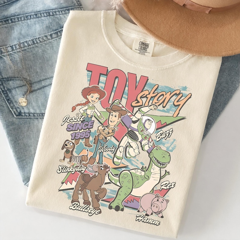 May include: Cream-colored t-shirt featuring the words "Toy Story" in a retro font, along with illustrations of characters like Woody, Buzz Lightyear, and Jessie. The shirt also includes the text "Jessie Since 1995".