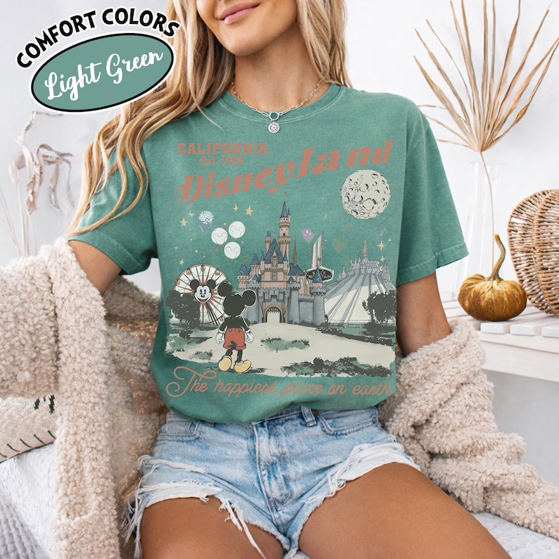 May include: Light green t-shirt with a Disneyland graphic. The graphic features Mickey Mouse, a castle, and the text "Disneyland" and "The happiest place on earth". The shirt has the words "Comfort Colors" and "Light Green" in a circle.
