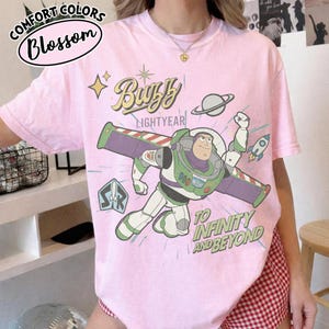 Może przedstawiać: Jasnor&oacute;żowa koszulka z grafiką Buzza Astrala z Toy Story. Projekt zawiera tekst "Buzz Lightyear" i "To Infinity and Beyond". Koszulka ma r&oacute;wnież tekst "Comfort Colors Blossom" w okrągłym wzorze.
