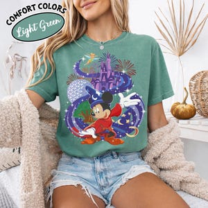 Może przedstawiać: Jasnozielona koszulka z grafiką Myszki Miki w stroju czarodzieja, z zamkiem, fajerwerkami i innymi elementami. Koszulka ma napisy "Comfort Colors" i "Light Green" w okrągłym wzorze.