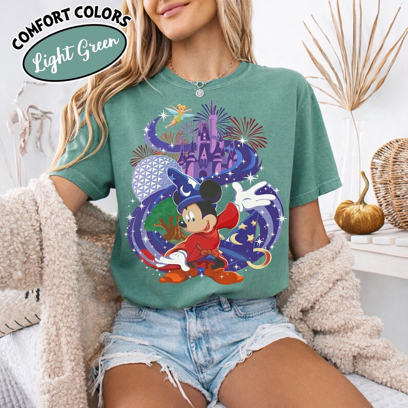 Może przedstawiać: Jasnozielona koszulka z grafiką Myszki Miki w stroju czarodzieja, z zamkiem, fajerwerkami i innymi elementami. Koszulka ma napisy "Comfort Colors" i "Light Green" w okrągłym wzorze.