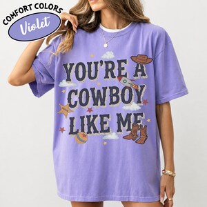 May include: A violet short-sleeve t-shirt featuring the text "YOU'RE A COWBOY LIKE ME" in dark blue letters. The graphic includes illustrations of a brown cowboy hat, a rocket ship, a sheriff star, a ball, and brown cowboy boots with clouds.