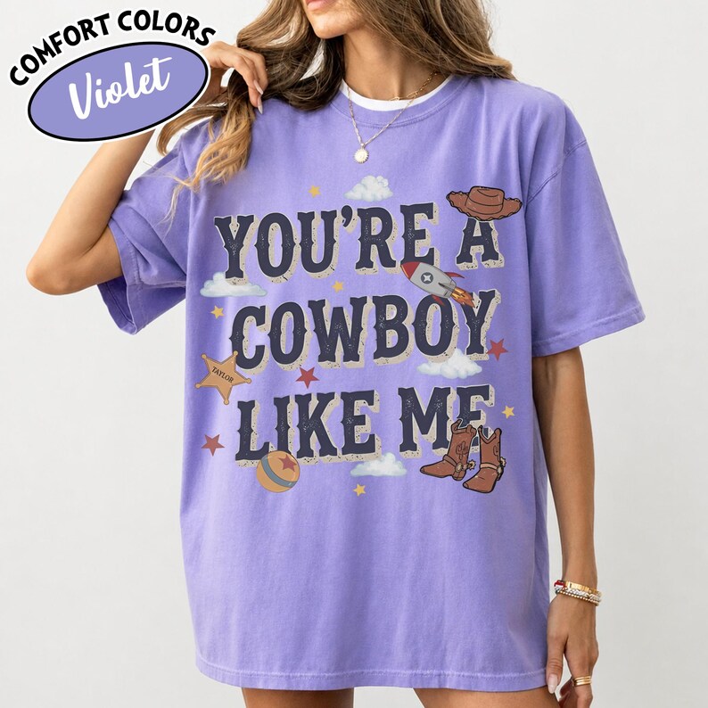 May include: A violet short-sleeve t-shirt featuring the text "YOU'RE A COWBOY LIKE ME" in dark blue letters. The graphic includes illustrations of a brown cowboy hat, a rocket ship, a sheriff star, a ball, and brown cowboy boots with clouds.