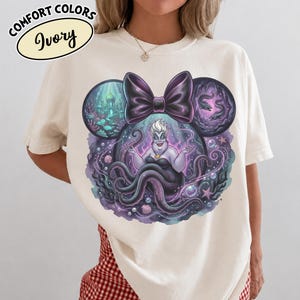 May include: Ivory-colored t-shirt featuring a graphic of Ursula from The Little Mermaid, within a Mickey Mouse ear design. The design includes a purple bow, sea creatures, and the text "Comfort Colors Ivory".