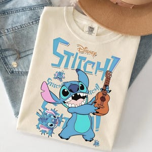 Pu&ograve; includere: T-shirt color crema con il personaggio Disney Stitch che suona un ukulele. La maglietta ha la scritta "Stitch!" in blu, insieme a illustrazioni pi&ugrave; piccole di Stitch. Un cappello marrone e jeans blu sullo sfondo.