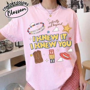 May include: A light pink short-sleeve t-shirt featuring a graphic print with the text "I KNEW IT I KNEW YOU", "infinity and beyond", and "YOU'VE GOT A FRIEND IN ME". The design includes illustrations of a beaded bracelet, a star, cowboy boots, blocks, and a hat.