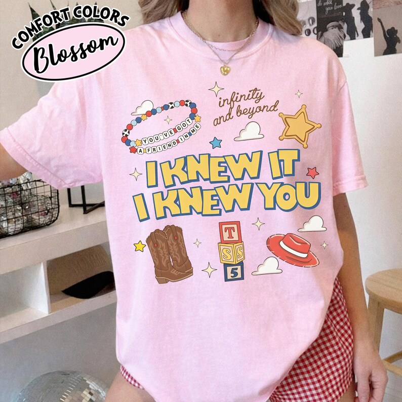 May include: A light pink short-sleeve t-shirt featuring a graphic print with the text "I KNEW IT I KNEW YOU", "infinity and beyond", and "YOU'VE GOT A FRIEND IN ME". The design includes illustrations of a beaded bracelet, a star, cowboy boots, blocks, and a hat.