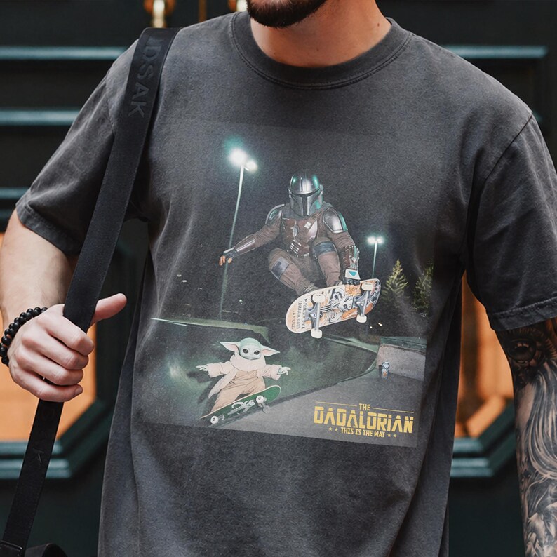 May include: Dark gray t-shirt featuring a graphic of The Mandalorian and Grogu skateboarding. The design includes the text "The Dadalorian" and "This is the way." The shirt has a vintage, distressed look.