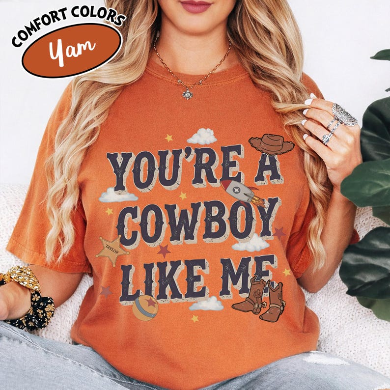 May include: A burnt orange short-sleeve t-shirt featuring the text "YOU'RE A COWBOY LIKE ME" in dark blue letters. The design includes graphics of a cowboy hat, a rocket, a ball, and cowboy boots, along with small stars and clouds.