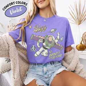 Może przedstawiać: Fioletowa koszulka Comfort Colors z grafiką Buzza Astrala i tekstem "Buzz Lightyear" i "To Infinity and Beyond". Koszulka jest w jasnofioletowym kolorze.