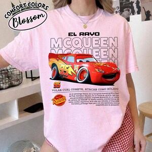 May include: Pink Comfort Colors t-shirt featuring a graphic of Lightning McQueen from the movie Cars. The design includes the text "EL RAYO MCQUEEN" and "95". The shirt also has the Rust-eze logo.