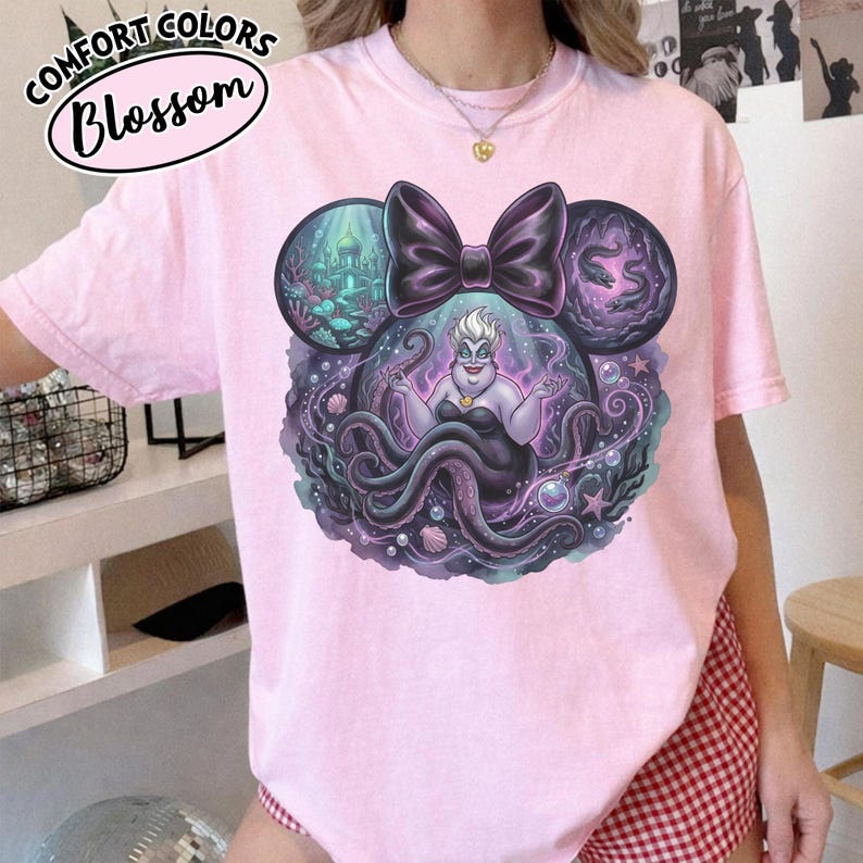 May include: Light pink t-shirt featuring a graphic of Ursula from The Little Mermaid, with a black bow and Mickey Mouse ears design. The graphic includes an underwater scene and the text "Comfort Colors Blossom".