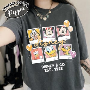 Op de afbeelding: Een donkergrijs Comfort Colors T-shirt met een Disney-ontwerp. Het ontwerp bevat afbeeldingen van Goofy, Minnie, Mickey, Daisy, Donald en Pluto in polaroid-stijl frames. De tekst "Disney & Co EST. 1928" is eronder gedrukt.