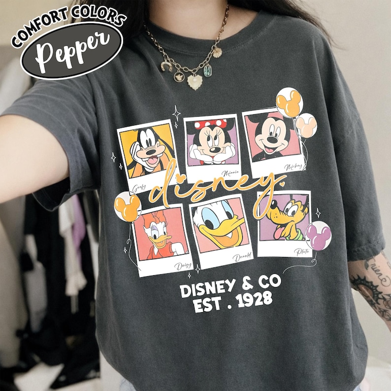 Op de afbeelding: Een donkergrijs Comfort Colors T-shirt met een Disney-ontwerp. Het ontwerp bevat afbeeldingen van Goofy, Minnie, Mickey, Daisy, Donald en Pluto in polaroid-stijl frames. De tekst "Disney & Co EST. 1928" is eronder gedrukt.
