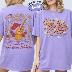 May include: A violet t-shirt featuring a graphic on the back and front. The back displays a cowboy hat, guitar, bull skull, and text reading Cowgirl Like Me and Takes One to Know One. The front features the text it's a Toy Story Baby just say yes.