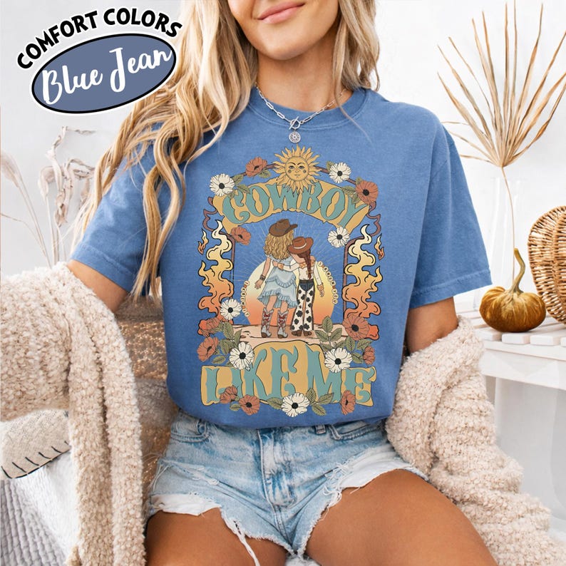 May include: A blue short-sleeve t-shirt featuring a graphic print of two figures in cowboy hats standing together. The text "COWBOY LIKE ME" is printed in large letters. The design includes a sun, flowers, and flame-like patterns in warm tones.