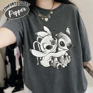 May include: Dark gray Comfort Colors Pepper t-shirt featuring a graphic of two cartoon characters hugging. The shirt has short sleeves and a crew neck. The text "COMFORT COLORS" and "Pepper" are printed on the top left of the shirt.