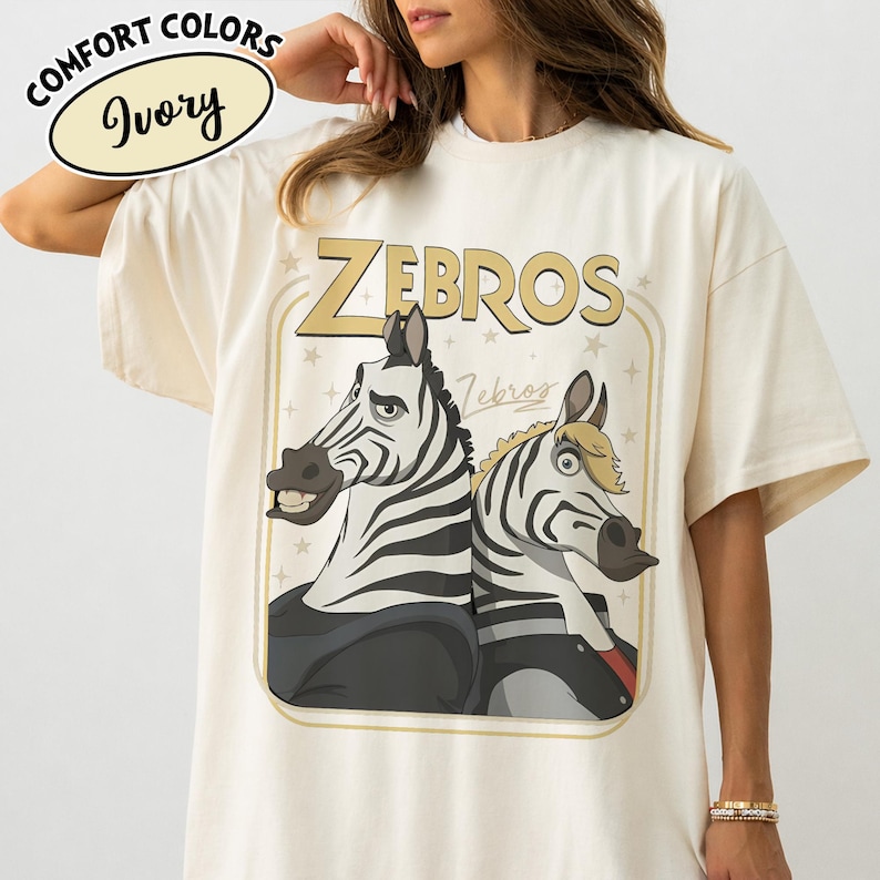 Puede incluir: Camiseta de color marfil con el texto "ZEBROS" en letras doradas. La camiseta presenta una ilustraci&oacute;n de dibujos animados de dos cebras. Las palabras "Comfort Colors" e "Ivory" est&aacute;n en un c&iacute;rculo en la parte superior izquierda.