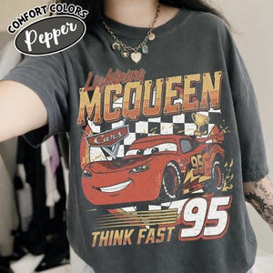 May include: A dark grey t-shirt with a graphic of Lightning McQueen from the Cars film. The design includes the text "Lightning McQueen", "95", and "Think Fast" in orange and white. The shirt has a vintage, distressed appearance.