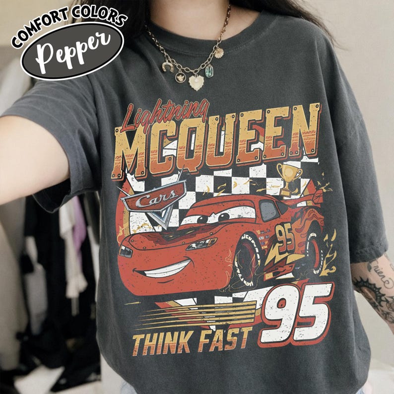 May include: A dark grey t-shirt with a graphic of Lightning McQueen from the Cars film. The design includes the text "Lightning McQueen", "95", and "Think Fast" in orange and white. The shirt has a vintage, distressed appearance.