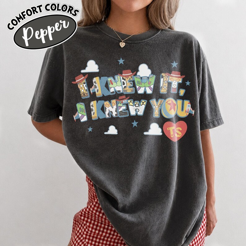 May include: A charcoal grey short-sleeve t-shirt featuring the text I KNEW IT, I KNEW YOU in a multi-colored font with character-themed patterns. The design includes small clouds, stars, and a red heart with the letters TS in the bottom right corner.
