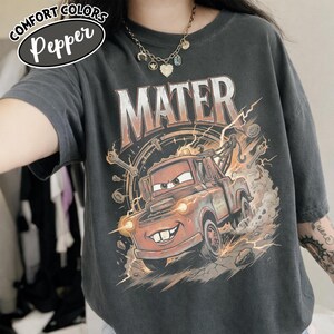 Puede incluir: Camiseta gris oscuro con un dise&ntilde;o de Mate de la pel&iacute;cula Cars. El gr&aacute;fico muestra a Mate con la palabra "MATER" encima, rodeado de herramientas y rayos. La camiseta tambi&eacute;n tiene las palabras "COMFORT COLORS" y "Pepper".