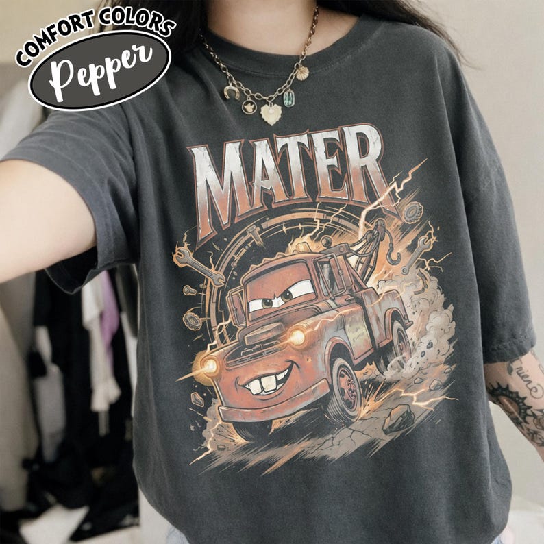 Puede incluir: Camiseta gris oscuro con un dise&ntilde;o de Mate de la pel&iacute;cula Cars. El gr&aacute;fico muestra a Mate con la palabra "MATER" encima, rodeado de herramientas y rayos. La camiseta tambi&eacute;n tiene las palabras "COMFORT COLORS" y "Pepper".