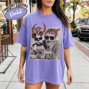 May include: Violet Comfort Colors t-shirt featuring a sepia-toned image of Walt Disney and Mickey Mouse wearing sunglasses. The word "WALT" is printed above the image in a burgundy color. The shirt is a solid violet color.