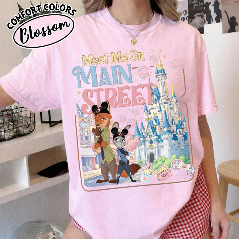 May include: Pink t-shirt with the text "Meet Me On Main Street" and a graphic featuring cartoon characters and a castle. The shirt also has the words "Comfort Colors" and "Blossom" printed on it.