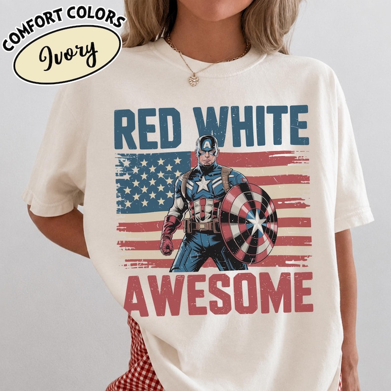 K&ouml;nnte beinhalten: Elfenbeinfarbenes T-Shirt mit einer Grafik von Captain America und dem Text "RED WHITE AWESOME". Das Design beinhaltet eine verblasste amerikanische Flagge. Das Shirt hat auch die Worte "COMFORT COLORS" und "Ivory" in einem Oval.