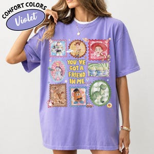 May include: Violet Comfort Colors t-shirt featuring a colorful graphic of Toy Story characters in postage stamp designs. The shirt includes the phrase "You've Got A Friend In Me" in yellow text. The shirt has short sleeves and a crew neck.