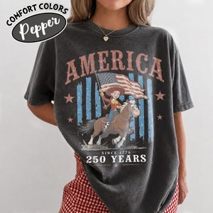 Puede incluir: Una camiseta gris carb&oacute;n con un estampado gr&aacute;fico que incluye el texto AMERICA en la parte superior y 250 YEARS SINCE 1776 en la inferior. El dise&ntilde;o muestra un personaje de dibujos animados montando un caballo marr&oacute;n con una bandera estadounidense.