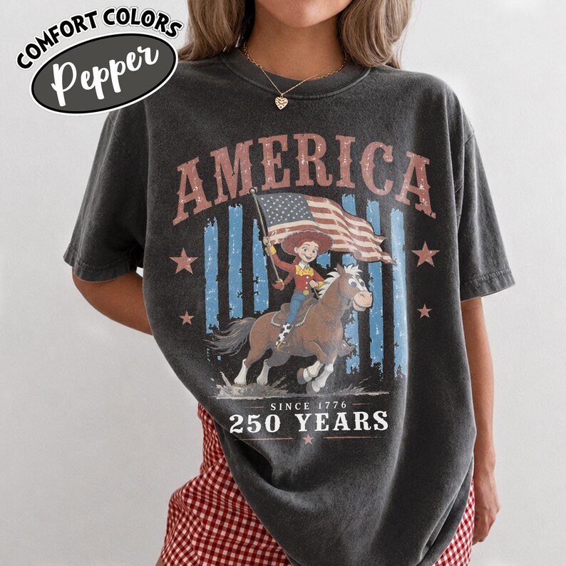 Puede incluir: Una camiseta gris carb&oacute;n con un estampado gr&aacute;fico que incluye el texto AMERICA en la parte superior y 250 YEARS SINCE 1776 en la inferior. El dise&ntilde;o muestra un personaje de dibujos animados montando un caballo marr&oacute;n con una bandera estadounidense.