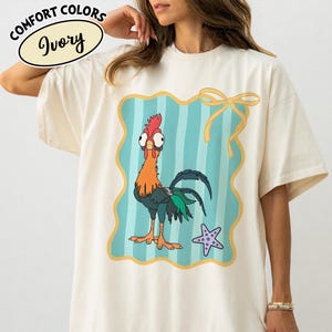May include: Ivory-coloured t-shirt with a cartoon rooster design inside a teal and gold frame. The rooster has orange, red, and green details. The shirt also has the words "COMFORT COLORS" and "Ivory" printed on it.